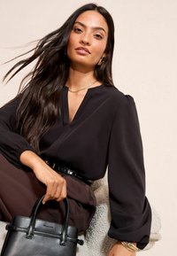 Hosszú, sötét hajú nő fekete blúzt és barna nadrágot visel, fekete kézitáska van nála, egy textúrázott széken ül egy semleges háttér előtt.