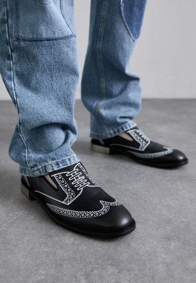 Person wearing light blue denim jeans and black leather dress shoes with white decorative stitching, standing on gray floor.