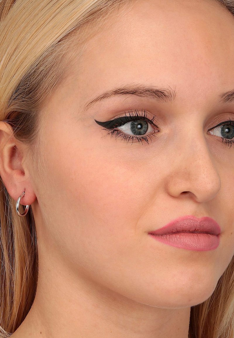 Primo piano di un volto con lunghi capelli biondi. I tratti includono eyeliner nero a forma di ala, labbra piene rosa e piccoli orecchini a cerchio in argento.