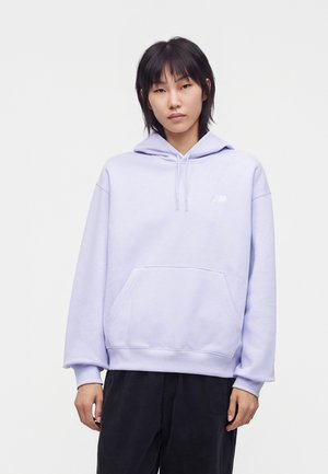 Jeune personne aux cheveux noirs raides, vêtue d'un sweat à capuche violet clair et d'un pantalon noir, debout devant un fond blanc uni.