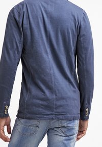 Uomo che indossa un blazer casual blu navy con polsini con bottoni e jeans blu sbiaditi, mostrato di spalle su uno sfondo bianco.