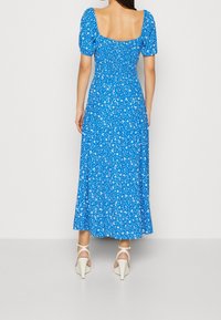 Niebieska kwiatowa sukienka midi z krótkimi bufkami, prostym dekoltem z tyłu i marszczonym materiałem. Noszona z beżowymi sandałami na wysokim obcasie.