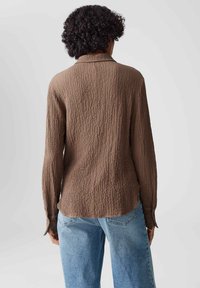 Braun strukturierte langärmlige Bluse mit lockerer Passform und Kragen, kombiniert mit hellblauen hochtaillierten Jeans, von hinten betrachtet.