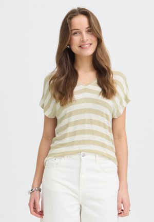 Femme souriante aux longs cheveux bruns portant un haut beige et blanc rayé à manches courtes et un pantalon blanc sur un fond uni.