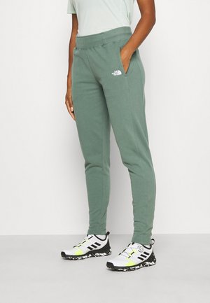 Person iført grønne North Face joggingbukser og hvide Adidas sneakers, stående mod en ensfarvet baggrund.