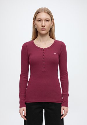 Femme aux cheveux blonds et raides portant une chemise henley bordeaux à manches longues et un pantalon noir, debout devant un fond blanc uni.