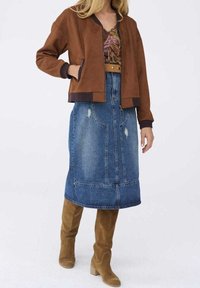 Femme portant une veste marron, un haut à motifs, une jupe midi en jean bleu et des bottes hautes en daim beige, debout devant un fond uni.