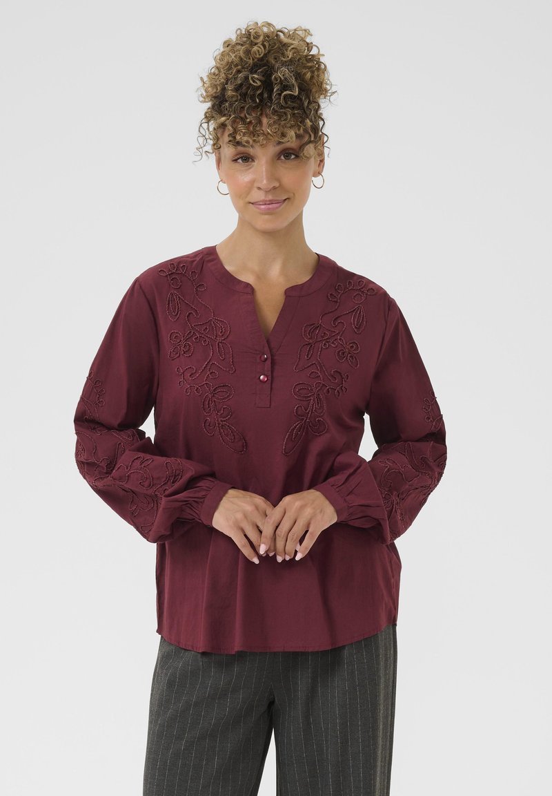 Bordeaux bluse med blomsterbroderi, tre-knap strop, og samlede manchetter, parret med gråstribede bukser.