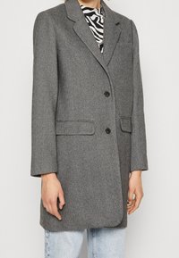 Blazer en espiga gris con solapas muesca, dos botones y bolsillos frontales. La tela tiene un acabado texturizado, combinado con una camisa a rayas.