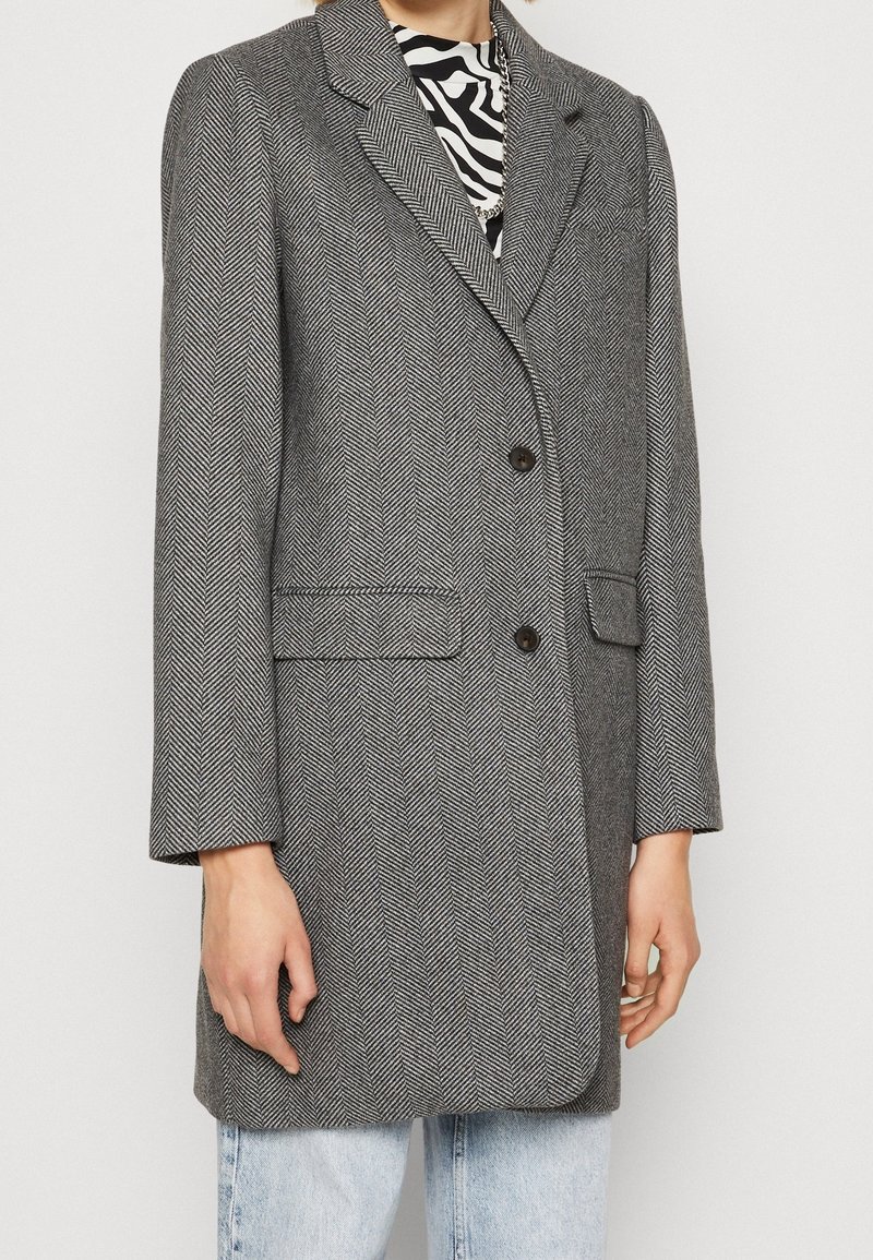 Blazer en espiga gris con solapas muesca, dos botones y bolsillos frontales. La tela tiene un acabado texturizado, combinado con una camisa a rayas.