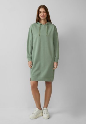 Helle grüne Kapuzenkleid aus weichem Material, mit Kordelzug an der Kapuze, seitlichen Schlitzen und langen Ärmeln, kombiniert mit weißen Sneakers.
