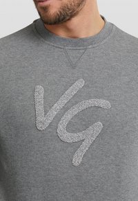 Felpa grigia con collo rotondo, caratterizzata da un logo "VG" testurizzato in un grigio più chiaro. Il tessuto appare morbido, con un taglio classico.