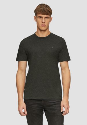 Jeune homme portant un t-shirt noir à motifs et un pantalon sombre, se tenant devant un fond gris clair uni.