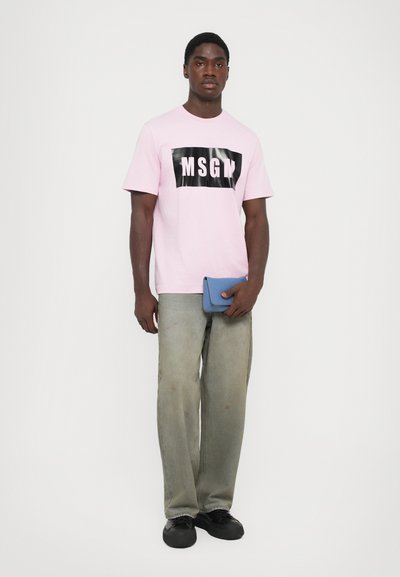 Lys rosa t-skjorte med en svart "MSGM" grafikk, parret med løse grå jeans og svarte sko. Subjektet holder en liten blå lommebok.