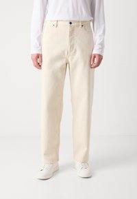 Un pantalon en coton couleur crème présente un design à jambe droite, des poches avant et un accent de bouton bleu. Porté avec des baskets blanches.