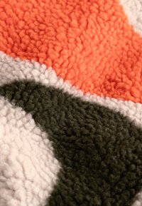 Fleece deka s abstraktním vzorem v oranžové, krémové a tmavě zelené barvě; texturovaný, načechraný povrch s různými výškami chlupů.