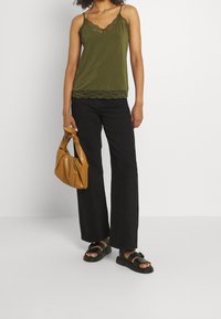Femme portant un haut sans manches en dentelle olive, un jean large noir, des sandales noires, tenant un sac à main en cuir beige à ses côtés.