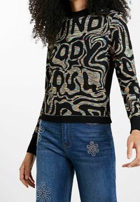 Pull en tricot avec un motif abstrait multicolore sur fond noir. Associé à un jean bleu orné de broderies florales sur les poches.
