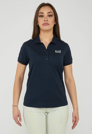 Donna con capelli lunghi e castani che indossa una polo blu navy EA7 e pantaloni chiari, in piedi davanti a uno sfondo chiaro e uniforme.