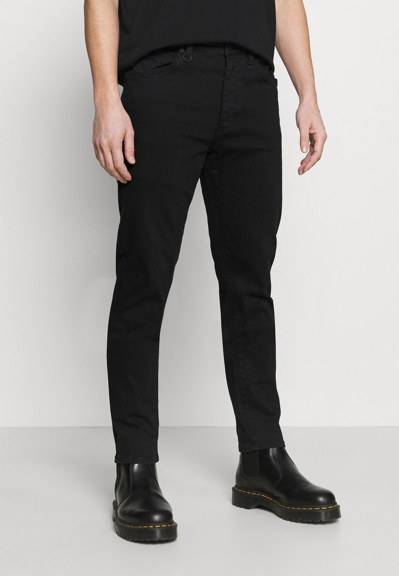 Man i svarta slim-fit byxor och svarta läder Chelsea-boots, står mot en slät ljus bakgrund.