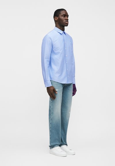 Ανοιχτό μπλε πουκάμισο με κουμπιά και μακριά μανίκια, με τσέπη στο μπροστινό μέρος, συνδυασμένο με ξεθωριασμένο μπλε τζιν και λευκά αθλητικά παπούτσια. Απλός, καθαρός σχεδιασμός.