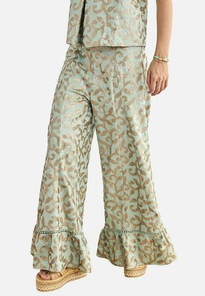 Pantalones de pierna ancha con patrón de hojas beige, bajos con volantes, combinados con sandalias de plataforma y una pulsera turquesa en la muñeca derecha.