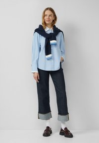 Lichtblauw gestreept overhemd met knopen, marineblauwe gebreide trui om de nek geknoopt, donkere denim cropped broek met opgerolde mouwen, bruine loafers.