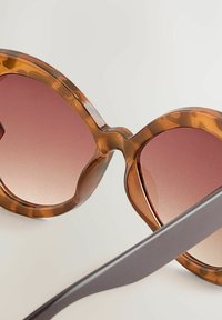 Lunettes de soleil en écaille de tortue avec une monture ronde surdimensionnée et des verres dégradés roses. Les branches sont marron foncé et ont un design épuré.