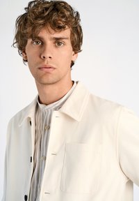 Vit lättviktsjacka med krage och knappstängning, som har en framficka och är parat med en randig skjorta under.