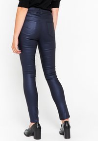 Tummansininen, korkeavyötäröinen leggings-malli, jossa on kiiltävä ja tekstuurinen finiš ja takataskut. Yhdistetty mustiin paksupohjaisiin kenkiin.