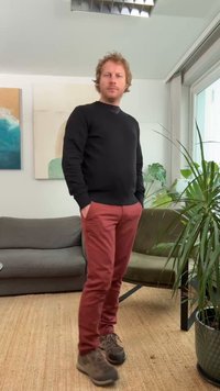 Fekete pulóver, piros nadrág és barna cipő. A szoba zöld növényt, szürke kanapét és absztrakt faliképet tartalmaz, világos környezetben.