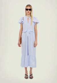 Vestido azul claro, até ao joelho, com mangas curtas com frufru, cintura com laço, decote em forma de olho de fechadura e bainha em camadas. Usado com sandálias pretas.