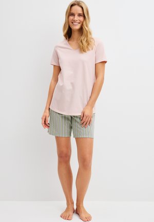 Donna sorridente, indossa una maglietta rosa pallido a maniche corte e pantaloncini a righe verticali verdi e bianchi, in piedi scalza davanti a uno sfondo bianco.