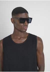 Occhiali da sole neri oversize con lenti piatte e montatura angolare. Indossati con una canotta nera a maniche corte dalla texture. Design semplice con accenti minimi.