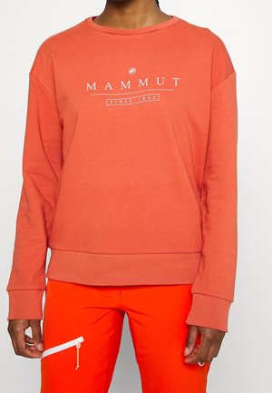 Personne portant un sweat-shirt à manches longues coraline de la marque Mammut et un pantalon orange vif avec une poche zippée sur la cuisse.