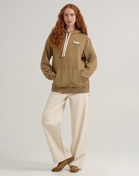 Felpa marrone con coulisse, caratterizzata da tasca frontale e logo. Abbinata a pantaloni cream larghi e scarpe slip-on marroni.