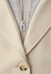 Beige blazer knapset over en grå lynlås-hoodie med en synlig sølv lynlås, der er centreret under blazerens krave.