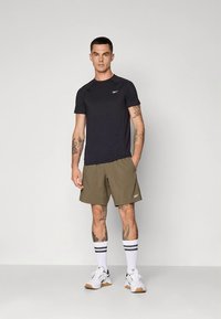 Černé atletické tričko s krátkými rukávy a olivově zelené šortky, k tomu bílé tenisky a pruhované ponožky, zdůrazňující přiléhavý střih a neutrální barvy.