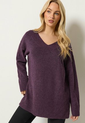 Blonde vrouw die een losse paarse V-hals trui en een zwarte broek draagt, staand tegen een effen lichte achtergrond, kijkt naar de zijkant.