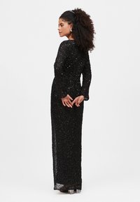 Abito lungo interamente ricoperto di paillettes nere, con maniche lunghe, superficie testurizzata e una silhouette aderente. Tacchi trasparenti completano il look.