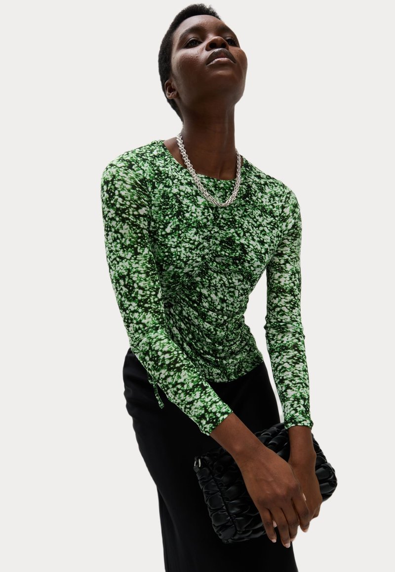 Groene blouse met lange mouwen en een gestructureerd bloemenpatroon. Gecombineerd met een zwarte, gewatteerde clutch. Strak aansluitende pasvorm met geplooide details bij de taille.