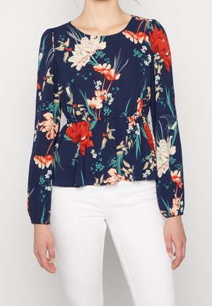 Vrouw die een donkerblauw bloesje met lange mouwen draagt met een rood-wit bloemmotief en een witte broek, getoond van hals tot heupen.