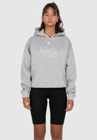 Grauer Hoodie mit einem weißen Frontgrafik-Logo, aus weichem Material gefertigt. Getragen mit schwarzen Radshorts, die einen lockeren Schnitt und lange Ärmel haben.