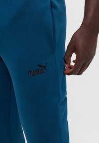 Κοντινό πλάνο μπλε φόρμας Puma με μαύρο λογότυπο κοντά στην τσέπη και ένα σκουρόχρωμο χέρι που ακουμπάει δίπλα στο πόδι.