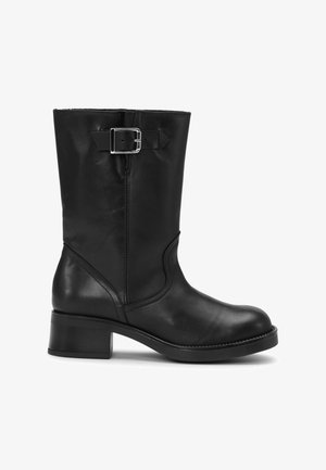 Bota negra de cuero hasta la mitad de la pantorrilla con tacón de bloque y una correa decorativa con hebilla plateada cerca de la parte superior, vista desde el lado exterior.