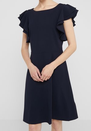 Mujer con vestido azul marino hasta la rodilla, mangas cortas con volantes en capas y escote redondo amplio, de pie con las manos entrelazadas al frente.