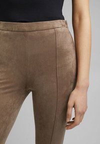 Close-up van een persoon in bruine suede leggings en een zwart top, met een zichtbare zijnaad en een ontspannen hand tegen het dijbeen tegen een grijze achtergrond.