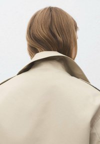Beige trenchcoat med en bred krage, som visar upp en slät yta. Bakifrån framhävs en skräddarsydd passform och en minimalistisk design.
