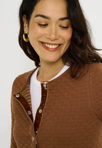 Cárdigan marrón acolchado con tela texturizada, cuello redondo y ribete de terciopelo contrastante con botones a presión y detalles dorados.