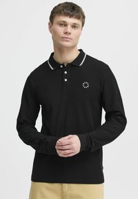 Crna dugorosna polo majica s bijelim obrubom na ovratniku, tri gumba na prednjoj strani i malim logotipom na lijevoj strani prsiju. Mekani materijal.